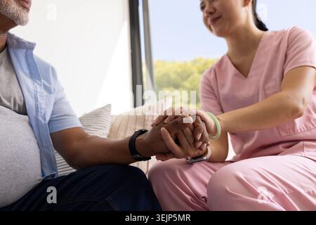 Infermiere e paziente diversi che tengono le mani sul divano a casa vicino alla finestra, infermiere che indossa scrub rosa Foto Stock