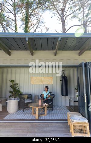 Donna africana adulta seduta su una sedia in rattan su una terrazza di legno con tavolino da caffè e borsa da pugno Foto Stock