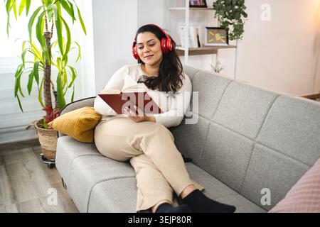 Donna con cuffie wireless che legge un libro su un divano Foto Stock