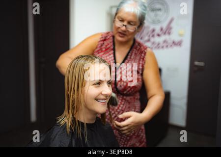 La parrucchiere taglia i capelli biondi bagnati del cliente al salone Foto Stock