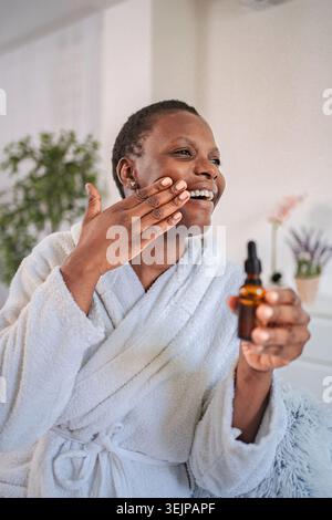 Donna sorridente che applica siero sul viso per la routine della cura della pelle Foto Stock