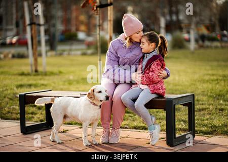 Madre e figlia sedute con il cane sulla panchina del parco Foto Stock
