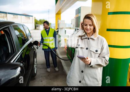 Donna che usa lo smartphone mentre fa rifornimento di carburante all'auto Foto Stock