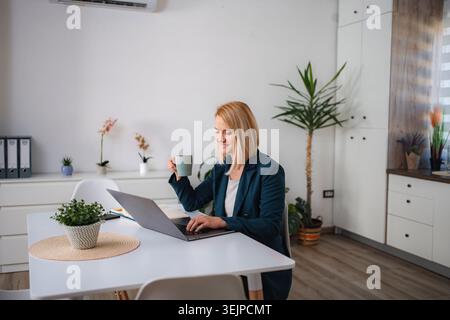 Donna sorridente che lavora da casa, usa un computer portatile e beve caffè Foto Stock