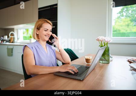 Donna sorridente che lavora al computer portatile mentre parla al telefono Foto Stock