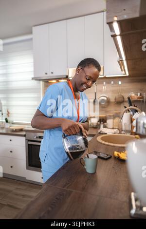 Infermiera sorridente che versa caffè nella sua cucina durante una pausa Foto Stock