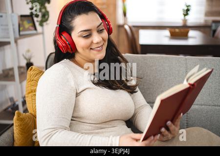 Donna con cuffie wireless che legge un libro su un divano Foto Stock