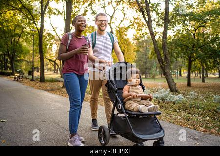 Una coppia gode di una piacevole passeggiata in un parco animato, circondato da foglie autunnali. Il loro bambino giocoso in un passeggino osserva la bella giornata, creando Foto Stock