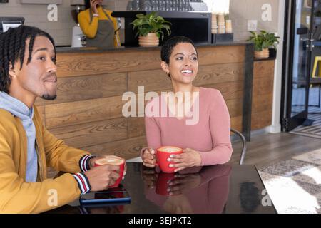 Coppia afroamericana seduta a un tavolo nero riflettente in un caffè con tazze rosse con smartphone. Caffè, latteart, accogliente, caldo Foto Stock
