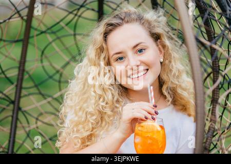 Adorabile donna positiva dai capelli leggeri gode delle vacanze estive, beve un cocktail d'arancia fresco mentre siede su una sedia sospesa, sorride Foto Stock