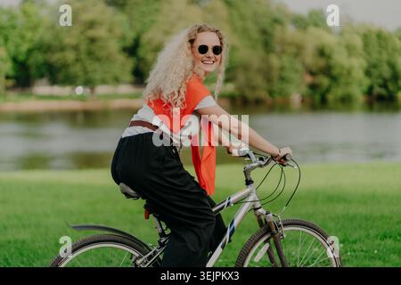 Foto di una giovane donna attraente con capelli ricci, giro in bicicletta, indossa occhiali da sole, maglietta, pantaloni, salta sulle spalle, si diverte Foto Stock