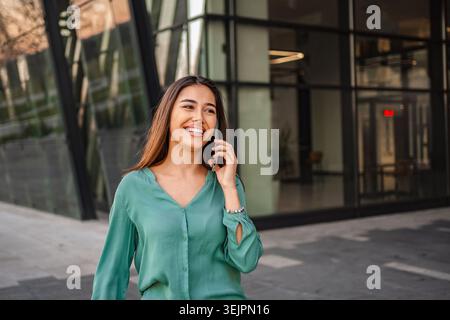 Giovane donna d'affari che parla al telefono e sorride vicino all'edificio degli uffici Foto Stock