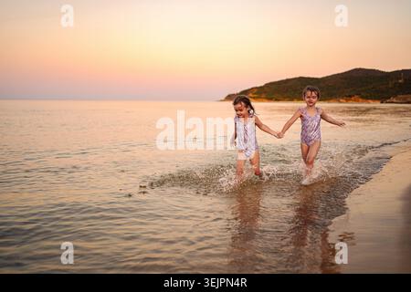 Le sorelle gemelle si godono una corsa gioiosa sulla riva al tramonto Foto Stock