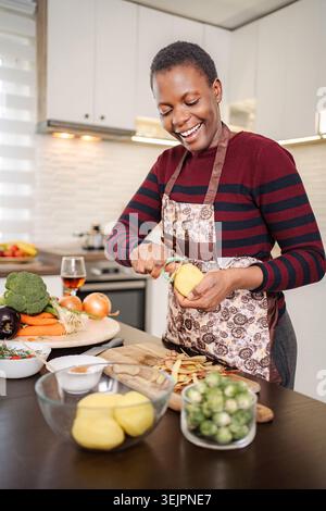 Giovane donna adulta felice che sbuccia le patate in cucina Foto Stock