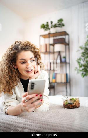 Giovane donna adulta che si rilassa sul letto utilizzando lo smartphone Foto Stock
