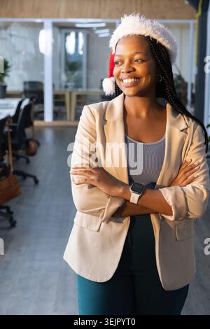 Donna afroamericana in piedi in un ufficio moderno con cappello, blazer e pantaloni color acqua che sorridono. Festa, posto di lavoro, manager, truffa Foto Stock