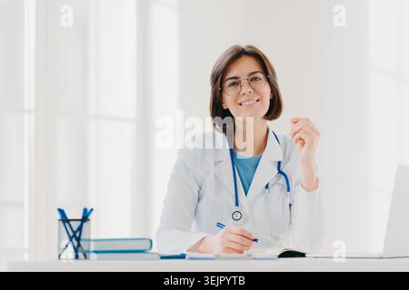 Giovane donna medico scrive su carta in clinica, creat prescrizione per il paziente, fa scartoffie in ospedale, indossa medico bianco g Foto Stock