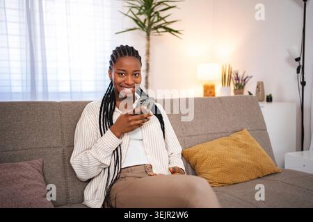 Giovane donna adulta che usa lo smartphone sorridente sul divano Foto Stock