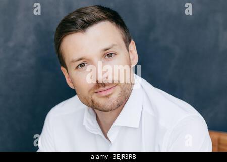 Foto isolata di un bel giovane lavoratore aziendale vestito con elegante camicia bianca, posa su sfondo scuro. Attraente Foto Stock