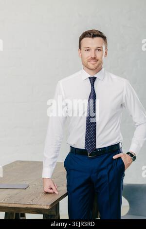 Foto di un bel lavoratore d'ufficio vestito con abiti formali, tiene una mano in tasca, l'altra sul tavolo, guarda con fiducia a c Foto Stock