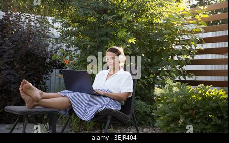 Donna di mezza età che indossa le cuffie con un laptop nel giardino vicino a casa in estate. Lavorare da qualsiasi luogo. Foto Stock