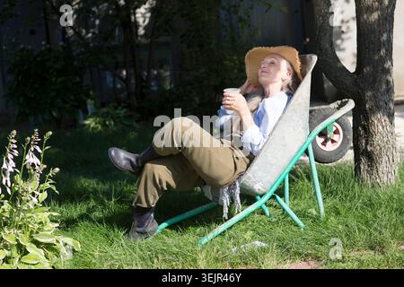 Una donna anziana con lunghi capelli grigi che indossa un cappello di paglia si siede in una carriola da giardino e beve tè nel giardino estivo vicino al Foto Stock