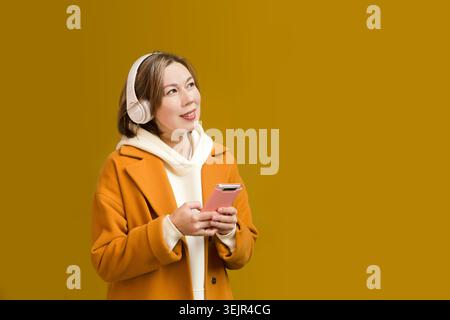 Una giovane donna con un cappotto giallo con le cuffie utilizza un telefono cellulare per strada della città sullo sfondo di un giallo Foto Stock