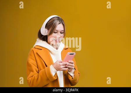 Una giovane donna con un cappotto giallo con le cuffie utilizza un telefono cellulare per strada della città sullo sfondo di un giallo Foto Stock