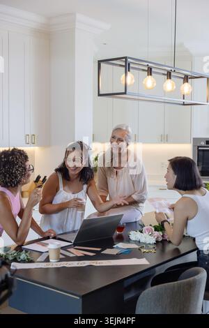 Donna anziana e tre donne adulte che si riuniscono a Kitchen Island, preparano carte con laptop e fiori. Multigenerazionale, collaborativo Foto Stock