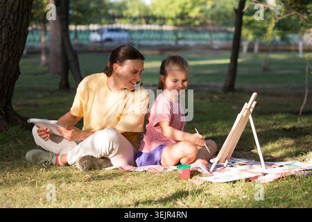 Famiglia felice mamma e figlia che si divertono a dipingere con un cavalletto in legno e a fare un picnic all'aperto nel parco cittadino sul green gra Foto Stock