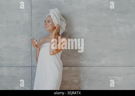 La giovane donna sorridente e premurosa applica fondamenti o idratanti sul viso, contiene una bottiglia di lozione per il corpo, avvolto in un asciugamano da bagno, co Foto Stock