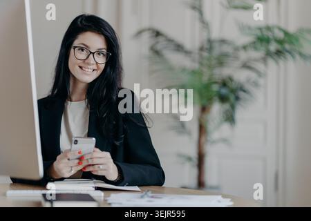 Una donna felice in ufficio con l'elegante giacca nera sfoglia lo smartphone con un'espressione allegra, invia messaggi di testo e lavora su im Foto Stock