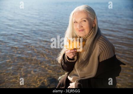 Una donna anziana con i capelli grigi che indossa un cappotto marrone tiene in mano una foglia d'acero giallo sulla riva del fiume. Vita lenta. IG Foto Stock