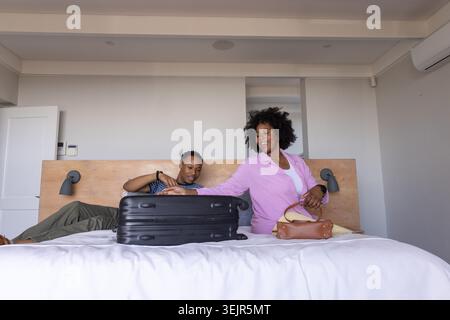 Coppia afroamericana seduta sul letto, che porta la valigia e che tiene una borsa di pelle in camera da letto. Bagagli, hotel, duo, viaggi, organizzazione Foto Stock