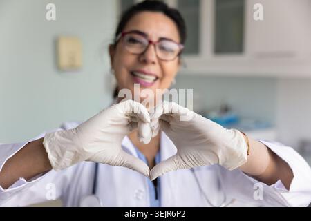Donna medica matura positiva che indossa guanti in lattice che fanno la mano cuore Foto Stock