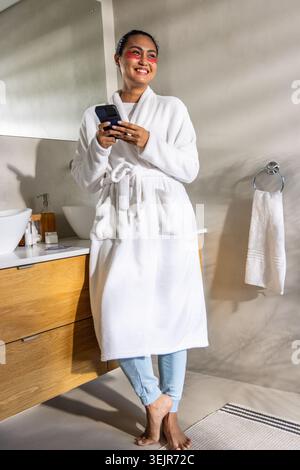 Donna asiatica adulta in piedi in bagno che tiene lo smartphone sorridente, indossa l'abito sopra i jeans arrotolati. Lavandino, specchio, asciugamano, Foto Stock