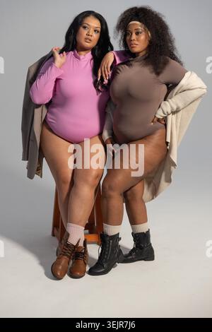 Posa in studio, diverse modelle femminili che indossano tute, cappotti, stivali, una seduta su sgabello. Duo, fashion, editoriale, onepiece Foto Stock