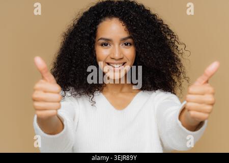 Giovane donna sorridente che mostra un gesto con i pollici sullo sfondo beige dello studio Foto Stock