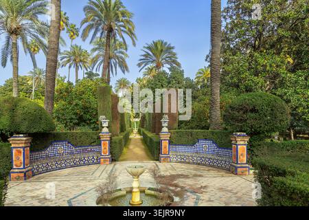 Vicolo decorativo nei giardini dell'Alcazar di Siviglia, Spagna, con una piccola fontana. Panchine piastrellate in ceramica sotto il cielo azzurro Foto Stock