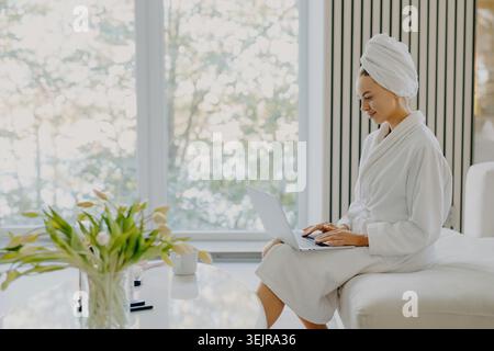 Donna in accappatoio che utilizza un computer portatile a casa dopo la doccia Foto Stock