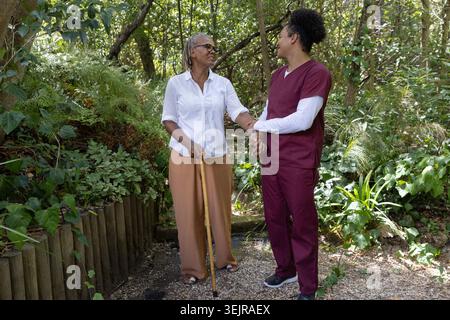 Donna anziana che tiene in mano canne di legno e caregiver in scrub di colore marrone di sostegno, in piedi su un sentiero di ghiaia. Cura dell'anziano, custode, vite umane Foto Stock