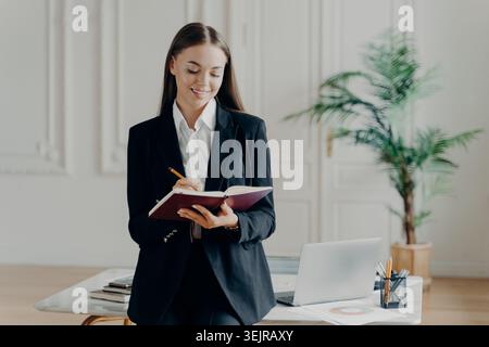 Donna d'affari che prende appunti in ufficio in piedi accanto alla scrivania con un computer portatile Foto Stock