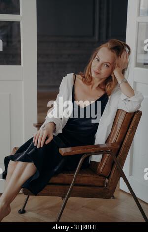 Donna dai capelli rossi seduta su una sedia di legno a casa con abito nero e camicia bianca Foto Stock