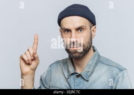 Uomo che punta le dita in verticale Studio con camicia in denim e berretto blu su sfondo grigio Foto Stock