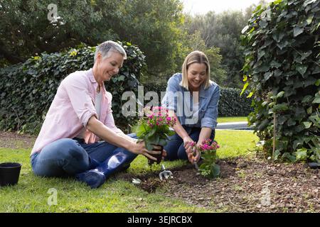 Mamma e figlia adulta si inginocchiano nel cortile di casa piantando fiori rosa con cazzuola, indossando denim. Giardinaggio, coltivazione, flo Foto Stock