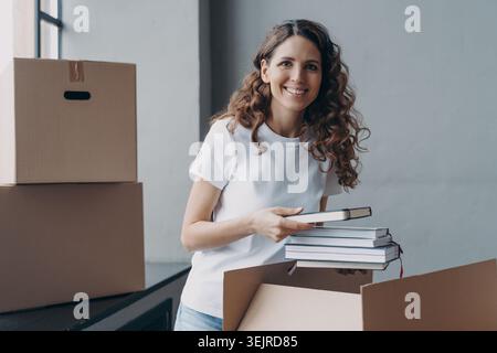 Sorridente giovane donna che imballa i libri nella scatola di cartone durante il trasferimento a casa e il trasferimento degli appartamenti in interni Foto Stock