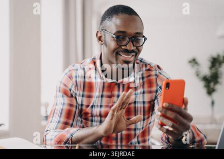 Happy Black Man effettua videochiamate sullo smartphone a casa e saluta Foto Stock