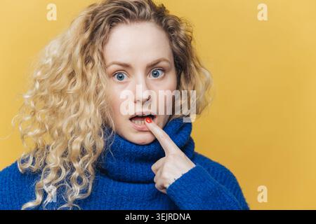 Donna bionda sorpresa che tocca le labbra con manicure rossa su sfondo giallo e indossa un pullover blu brillante in Studio Foto Stock