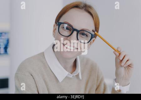 Donna sorridente e premurosa con gli occhiali che tengono la matita vicino alla testa in un ufficio luminoso per pianificare appunti di lavoro o studiare le idee in primo piano Foto Stock