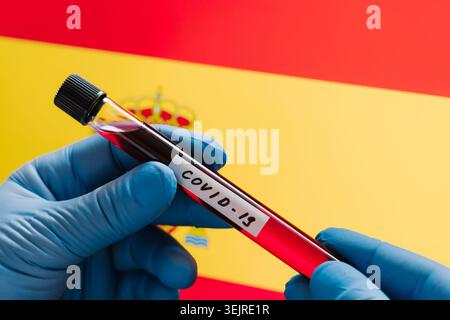 Primo piano di mani blu con guanti che tengono la provetta per test Covid con campione di sangue sopra la bandiera spagnola per le regole di screening Travel Health Foto Stock